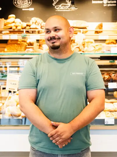 Eine Person steht vor einer gut bestückten Bäckereitheke mit verschiedenen Brotsorten und Backwaren. Im Hintergrund sind Regale mit frischen Backwaren und ein dekorativer Schriftzug zu sehen.