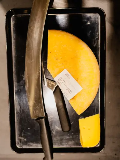 Ein großes Stück runder, gelber Käse liegt auf einem schwarzen Tablett. Daneben befinden sich ein Messer mit schwarzem Griff und ein weiteres kleineres Stück Käse. Auf dem Käse ist ein Etikett mit Informationen angebracht.