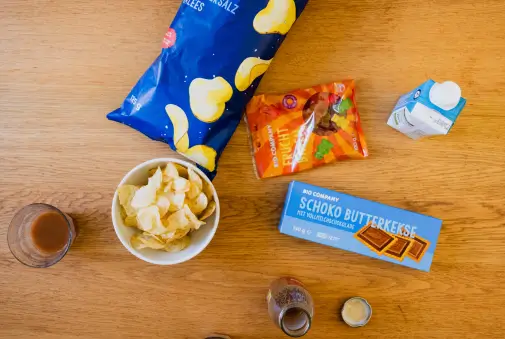 Eine Auswahl von Snacks und Getränken liegt auf einem Holztisch. Zu sehen sind eine geöffnete Chipstüte, eine Schüssel mit Chips, Schokokekse, eine Packung Gummibärchen, ein Getränkekarton und mehrere Gläser mit Getränken.