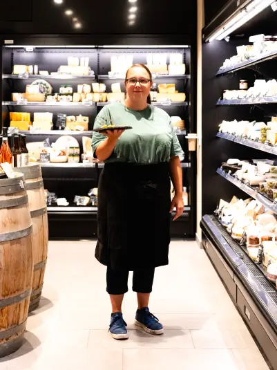 Eine Person in einem Supermarkt steht vor einer gut beleuchteten Käsetheke. Sie trägt ein grünes T-Shirt, eine schwarze Schürze und blaue Sneaker. In der Hand hält sie ein Tablett mit Käse. Links im Bild sind Holzfässer, rechts die Käseauslage.