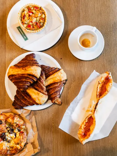  Eine Auswahl an To-Go-Speisen liegt auf einem Holztisch, darunter Croissants, ein belegtes Baguette, eine kleine Obsttarte und eine Pizza. Daneben steht eine Tasse Espresso.
