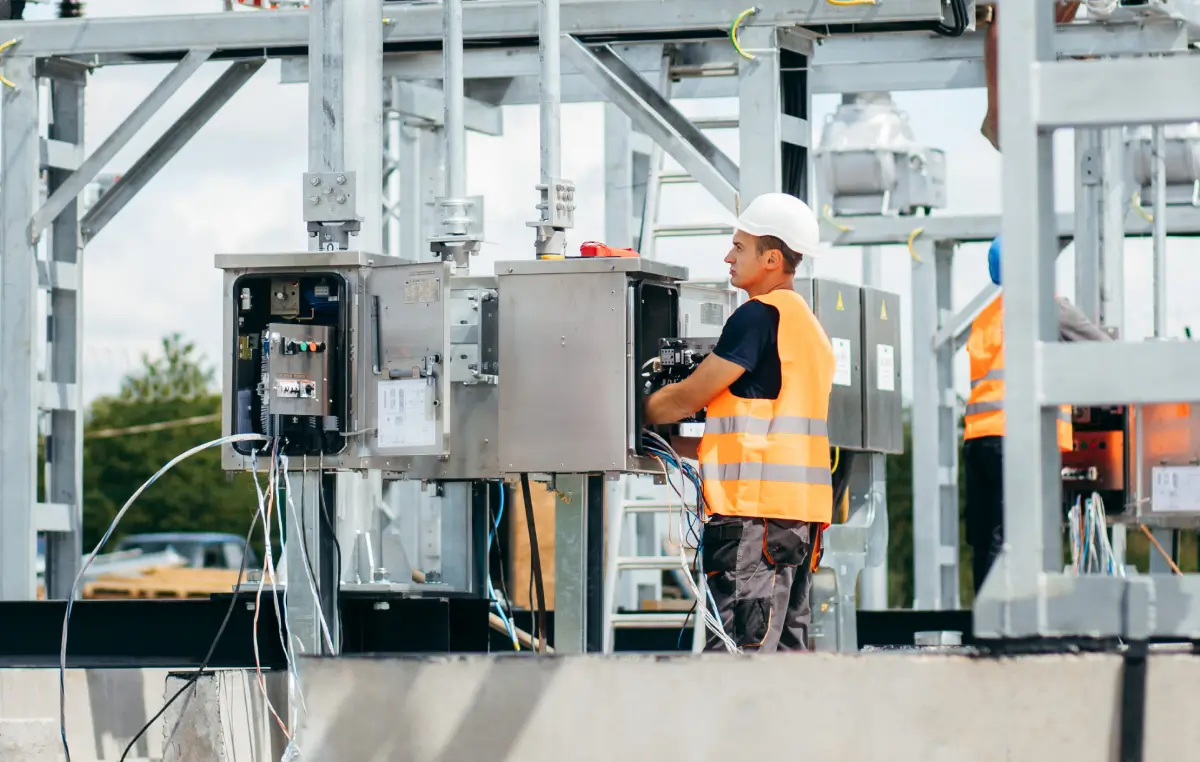Ein Arbeiter mit Helm und Warnweste arbeitet an einem offenen Schaltschrank auf einem Umspannwerksgelände mit Metallkonstruktionen und technischen Anlagen im Freien.