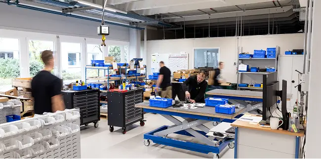 Mehrere Personen arbeiten und bewegen sich in einer modernen Werkstatt mit Werkbänken, Werkzeugen und Regalen.