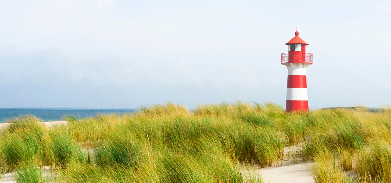 Ein rot-weiß gestreifter Leuchtturm steht hinter Dünen am Meer unter einem hellen Himmel.