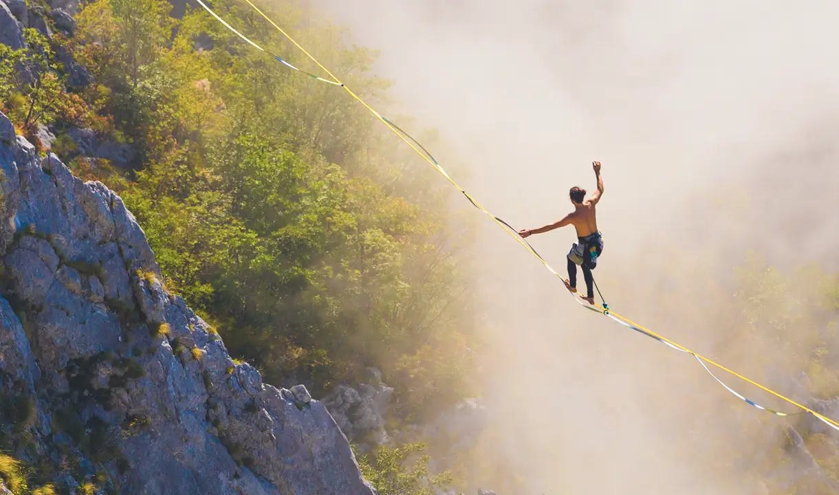Eine Person balanciert auf einer Slackline, die über eine Schlucht zwischen Felsen gespannt ist. Die Umgebung ist von grünen Bäumen und Felsen geprägt, und Nebel umhüllt die Szene teilweise. Die Person ist oberkörperfrei und trägt Kletterausrüstung.