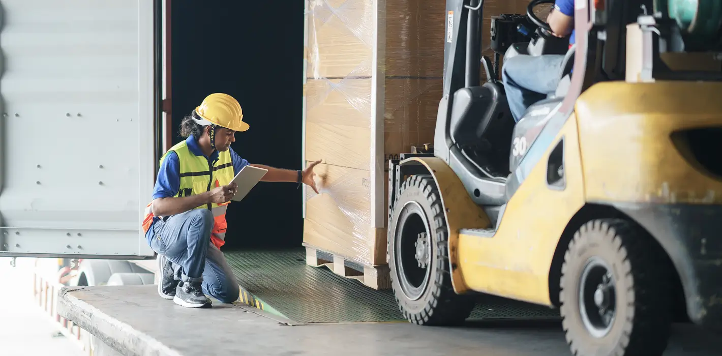 Ein Gabelstapler fährt eine Palette an, während ein Arbeiter mit Schutzweste und Helm die Lieferung überprüft.