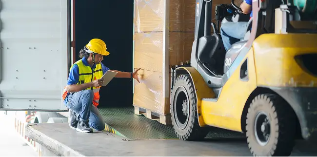 Ein Gabelstapler fährt eine Palette an, während ein Arbeiter mit Schutzweste und Helm die Lieferung überprüft.
