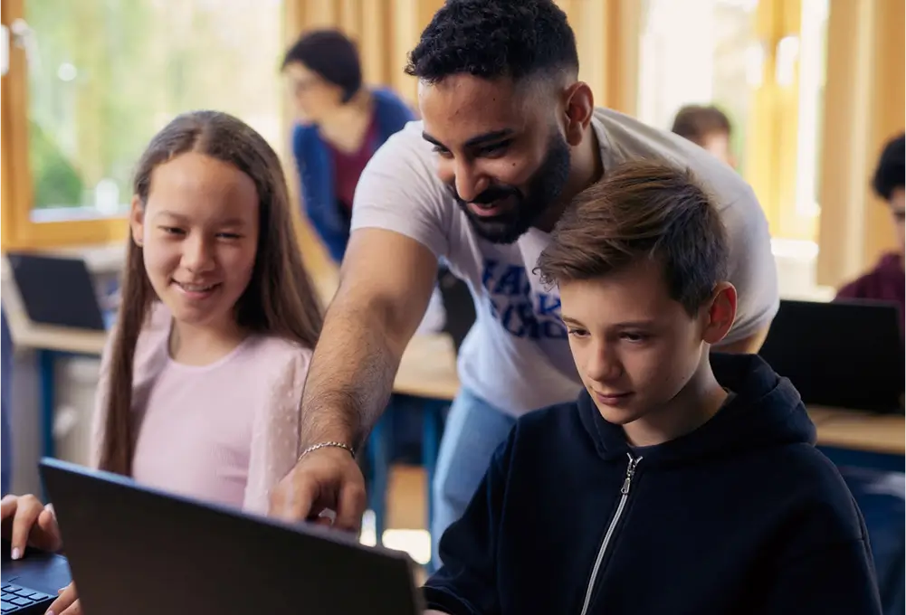 Ein Erwachsener unterstützt zwei Kinder beim Arbeiten an Laptops in einem modernen Klassenraum. Im Hintergrund sind weitere Kinder an Computern zu sehen.