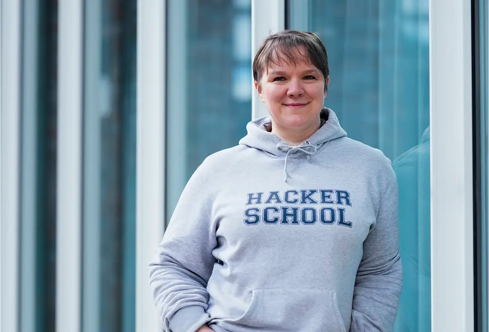 Eine Person steht vor einer Glasfassade und trägt einen grauen Hoodie mit der Aufschrift „Hacker School“.
