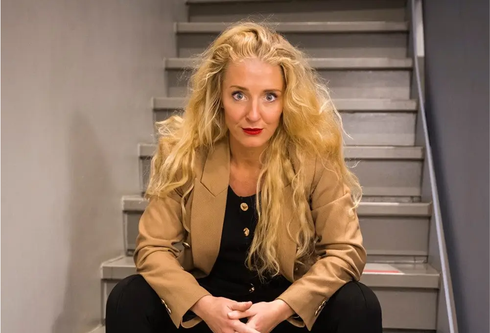 Eine Frau mit langen blonden Haaren sitzt in einem beigen Blazer auf einer Treppe in einem Innenraum.