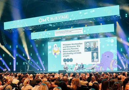 Eine große Bühne der „OMR Blue Stage“ mit modernem Design zeigt eine Podiumsdiskussion zum Thema „Millionen-Reichweiten und Shitstorms“. Im Hintergrund ist ein großer Bildschirm mit Illustrationen und Titelgrafiken, während ein dichtes Publikum aufmerksam zuhört.