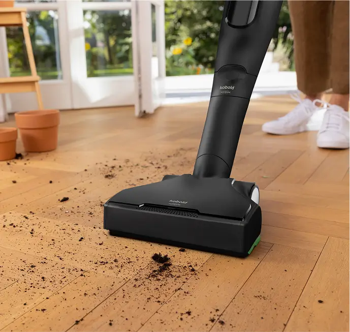 Ein Staubsauger entfernt Erde von einem Holzboden neben Blumentöpfen.