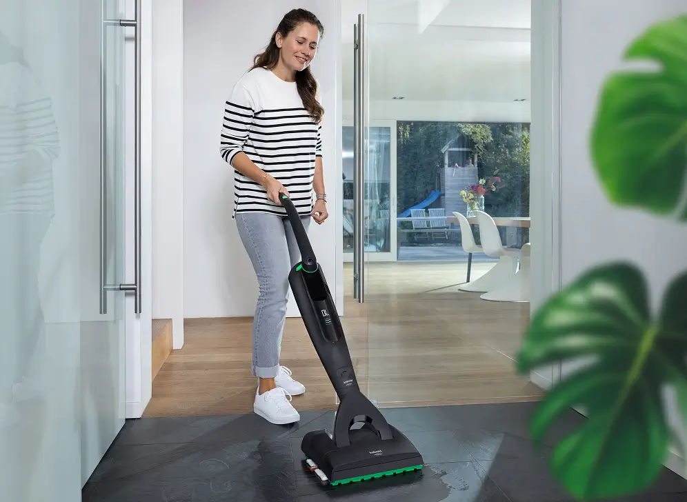 Eine Person reinigt mit einem modernen Vorwerk-Bodenreiniger einen dunklen Fliesenboden in einem hellen, modernen Wohnraum. Im Hintergrund ist ein Essbereich mit großen Fenstern und Blick in den Garten zu sehen.