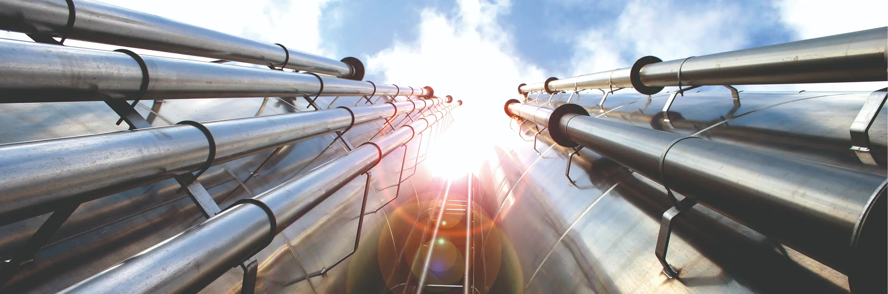 Blick nach oben auf mehrere große, glänzende Rohre mit Sonnenlicht im Hintergrund.