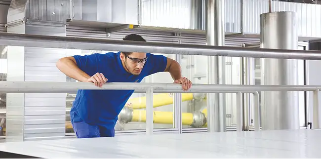 Arbeiter in blauer Kleidung beugt sich über ein Geländer in einer Produktionshalle.