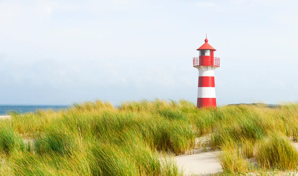 Ein rot-weiß gestreifter Leuchtturm steht hinter Dünen am Meer unter einem hellen Himmel.