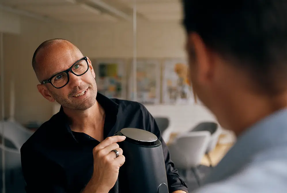 Eine Person zeigt einem Gegenüber ein rundes Detail an einem schwarzen Haushaltsgerät.