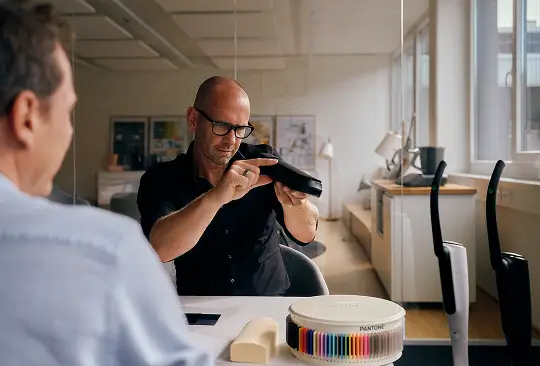 Zwei Personen sitzen an einem Tisch, eine prüft ein schwarzes Haushaltsgerät, daneben liegt ein Farbfächer.