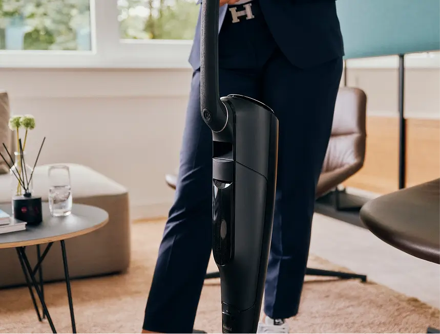Eine Person präsentiert einen schwarzen kabellosen Staubsauger im Stehen, im Hintergrund ein moderner Wohnraum.