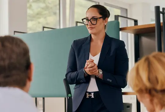 Eine Person im Anzug steht vor zwei sitzenden Personen und erklärt ein Produkt.