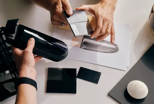 Zwei Personen besprechen Materialproben und ein schwarzes Gerät über einem technischen Entwurf.