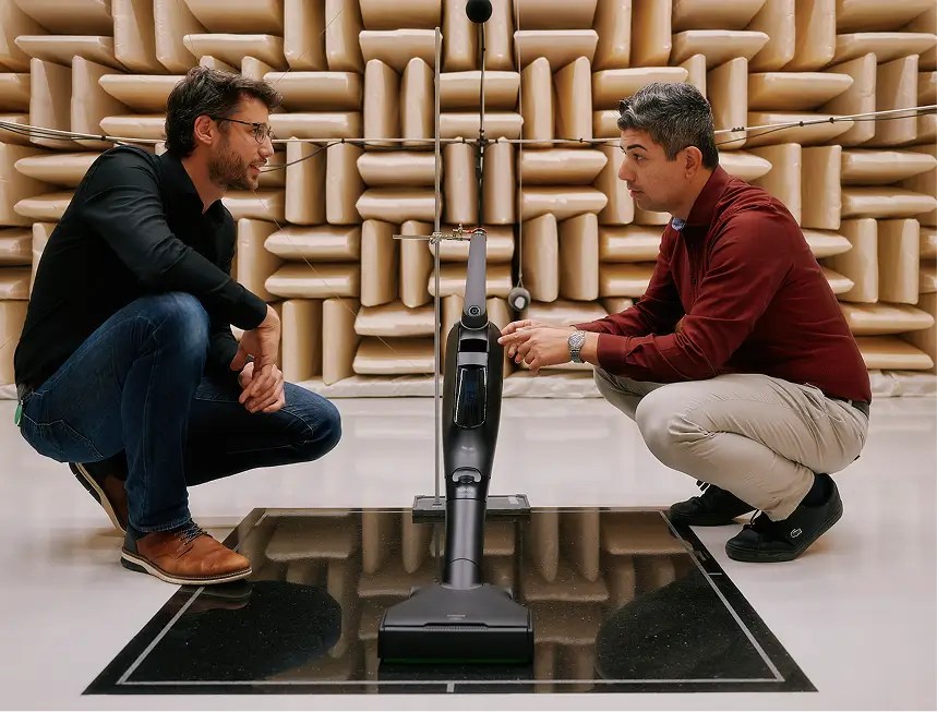 Zwei Männer hocken vor schwarzem Staubsaugerroboter auf Spiegelfläche in schallisoliertem Raum