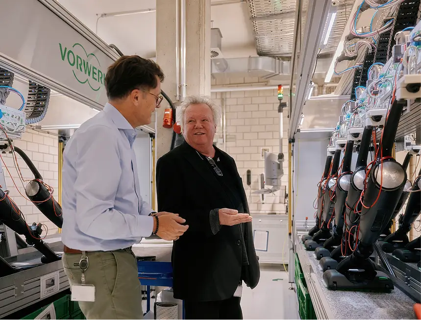 Zwei Männer im Gespräch vor einer Reihe verkabelter Staubsauger in Vorwerk-Testlabor