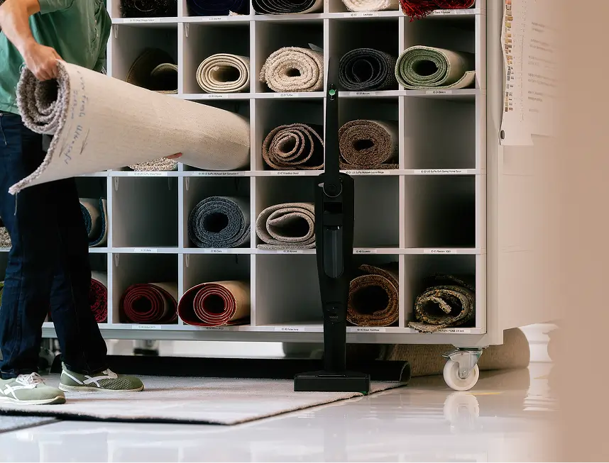 Mann stellt Teppichrolle vor Regal mit verschiedenen Teppichmustern neben schwarzem Staubsauger ab