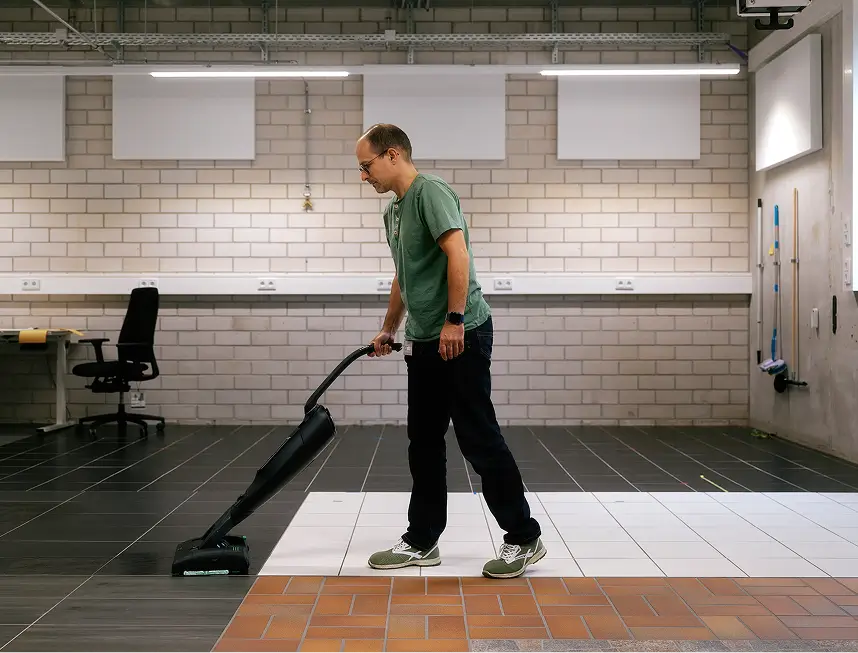 Mann mit grünem T-Shirt testet Staubsauger auf Fliesen und weißen Bodenplatten in Laborumgebung