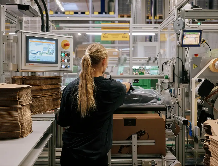 Mitarbeiterin mit Zopf bedient Verpackungsmaschine mit Touchscreen in Produktionshalle