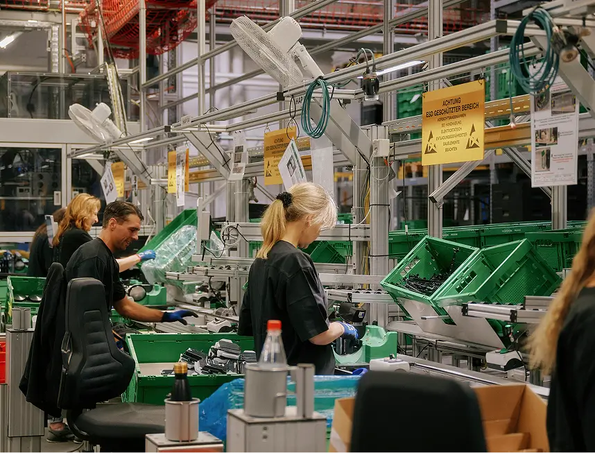 Arbeiter montieren VR-Headsets an Fließband mit grünen Kisten unter gelben Warnschildern