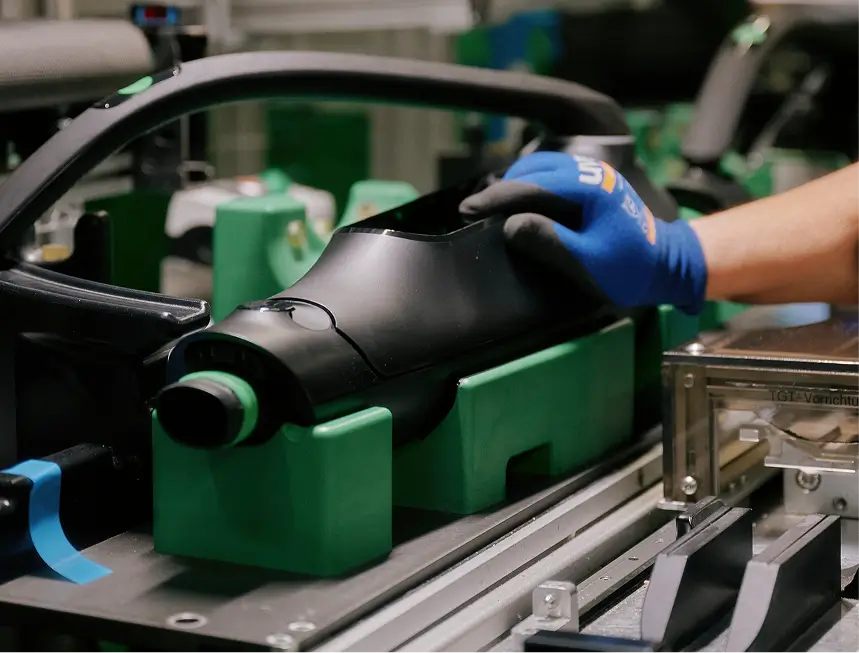 Hand in blauem Handschuh hält schwarzes VR-Headset über grüner Transportbox am Fließband