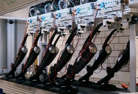 Mehrere schwarze Staubsauger mit Kabeln in automatisierter Testanlage vor Klinkerwand