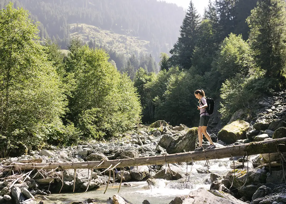 Eine Wanderin balanciert über einen Baumstamm, der über einen steinigen Gebirgsbach führt.
