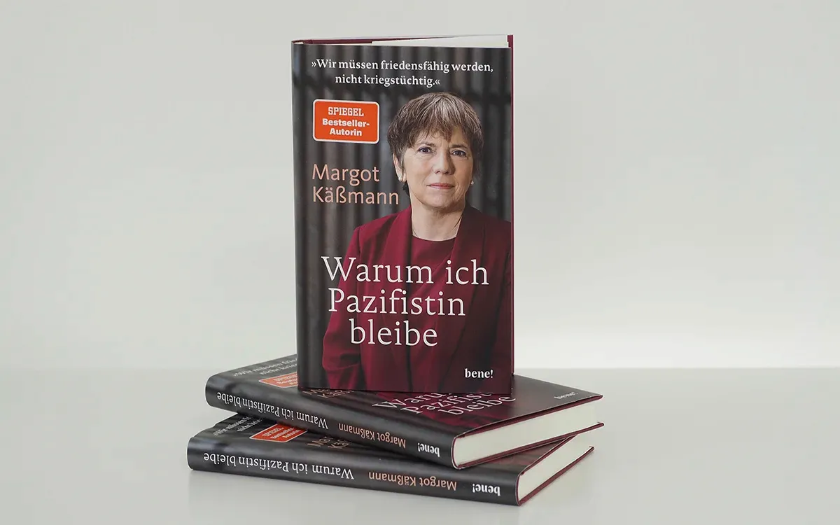 Das Buch „Warum ich Pazifistin bleibe“ steht aufrecht auf zwei liegenden Exemplaren desselben Buches. Auf dem Cover ist eine Frau in einem roten Oberteil abgebildet.