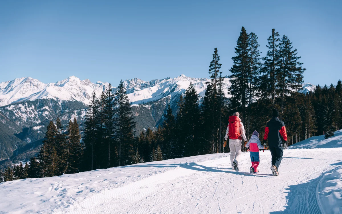 Zwei Erwachsene und ein Kind gehen auf einem verschneiten Weg zwischen Nadelbäumen und Bergen.