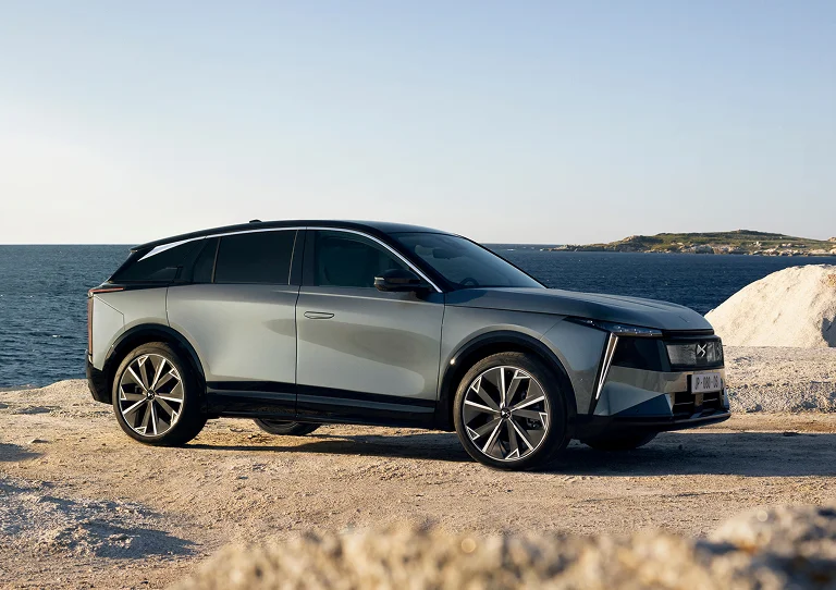Grauer SUV an einer felsigen Küste vor dunklem Meer und blauem Himmel.