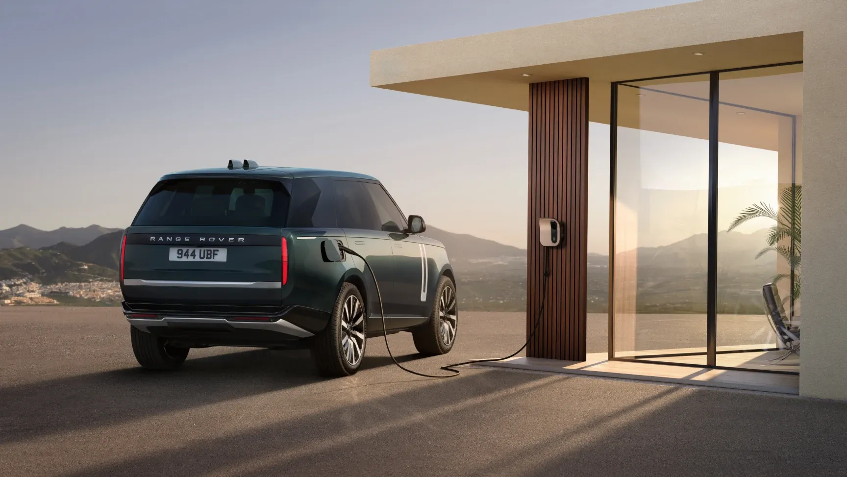 Ein dunkelgrüner SUV steht vor einem modernen Haus mit Glasfront und ist über ein Ladekabel an eine Wandladestation angeschlossen. Im Hintergrund sind Berge und ein klarer Himmel zu sehen.